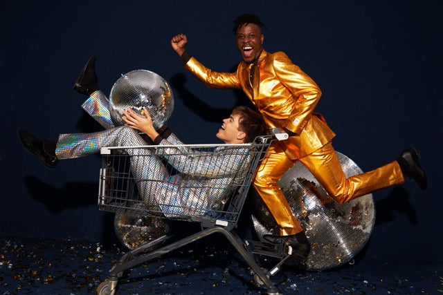 Two men celebrating, one laying in shopping cart and wearing discobal suit and one wearing golden men's suit